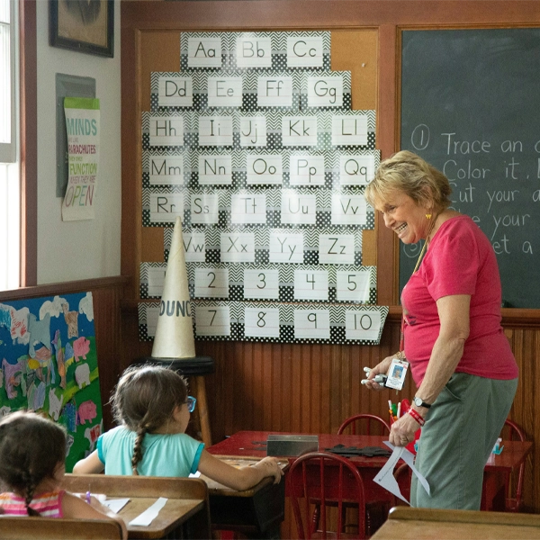 Blog Post 10.8.1 A teacher smiling while engaging with two young students in a classroom setting with alphabet and number posters on the wall